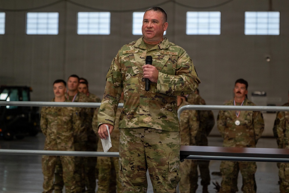 A U.S. Air Force servicemember identified by name tape as 'MCARDLE' is speaking into a microphone while holding papers in a military hangar setting. He is wearing Operational Camouflage Pattern (OCP) uniform with a 'U.S. AIR FORCE' tape visible on his chest. Several other uniformed personnel are visible in the background, with at least one wearing a medal.