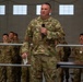 A U.S. Air Force servicemember identified by name tape as 'MCARDLE' is speaking into a microphone while holding papers in a military hangar setting. He is wearing Operational Camouflage Pattern (OCP) uniform with a 'U.S. AIR FORCE' tape visible on his chest. Several other uniformed personnel are visible in the background, with at least one wearing a medal.