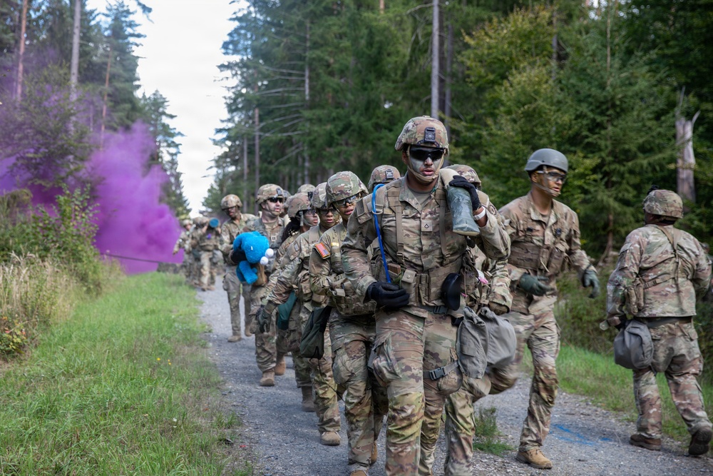 A column of soldiers in US Army MultiCam (OCP) uniforms marches along a forest trail during what appears to be a field training exercise. Purple and blue colored smoke grenades are visible in the background, commonly used as signals or markers during military exercises. Soldiers display face camouflage paint and carry various pieces of field equipment.