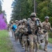 A column of soldiers in US Army MultiCam (OCP) uniforms marches along a forest trail during what appears to be a field training exercise. Purple and blue colored smoke grenades are visible in the background, commonly used as signals or markers during military exercises. Soldiers display face camouflage paint and carry various pieces of field equipment.