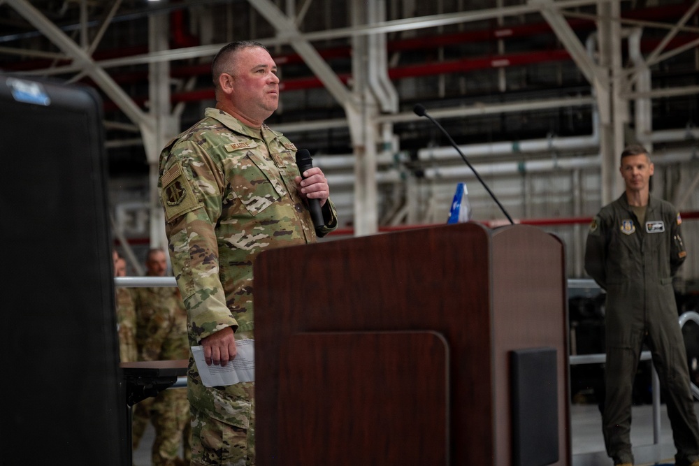 A military service member in OCP (Operational Camouflage Pattern) uniform is speaking at a podium while holding a microphone during what appears to be an official ceremony or briefing inside a military hangar. A second service member in an olive/sage green flight suit stands in the background to the right. The setting suggests a formal military event such as a change of command, award ceremony, or unit briefing.