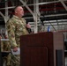 A military service member in OCP (Operational Camouflage Pattern) uniform is speaking at a podium while holding a microphone during what appears to be an official ceremony or briefing inside a military hangar. A second service member in an olive/sage green flight suit stands in the background to the right. The setting suggests a formal military event such as a change of command, award ceremony, or unit briefing.