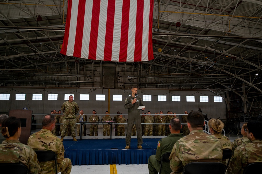 A military ceremony is taking place inside an aircraft hangar, with a large American flag hanging prominently from the ceiling. A service member in an olive green flight suit stands on a raised stage speaking into a microphone while holding papers, addressing an audience of seated and standing military personnel in camouflage uniforms. The formal setting, stage with blue skirting, and assembled troops suggest an official military event such as an award ceremony, change of command, or unit recognition.