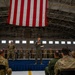 A military ceremony is taking place inside an aircraft hangar, with a large American flag hanging prominently from the ceiling. A service member in an olive green flight suit stands on a raised stage speaking into a microphone while holding papers, addressing an audience of seated and standing military personnel in camouflage uniforms. The formal setting, stage with blue skirting, and assembled troops suggest an official military event such as an award ceremony, change of command, or unit recognition.