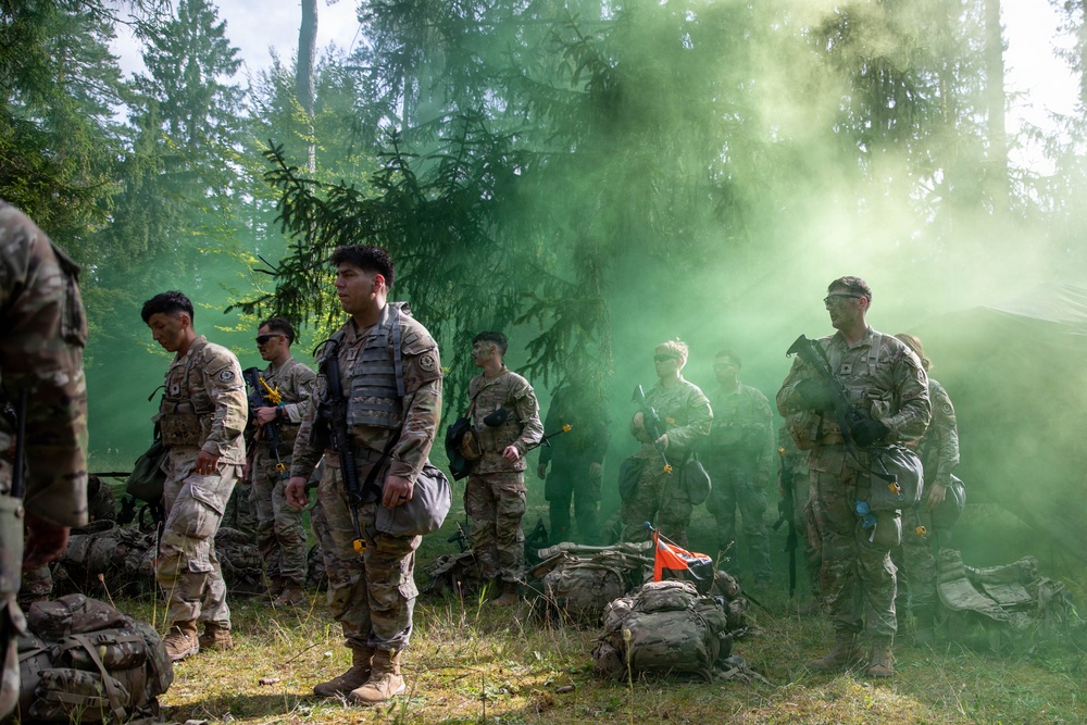 A group of military personnel in camouflage uniforms are assembled in a forested area amid a green smoke signal or grenade discharge. The soldiers are equipped with tactical gear, body armor, and weapons, suggesting a field training exercise or tactical operation. A small orange marker flag is visible on the ground among scattered rucksacks and equipment.