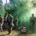 A group of military personnel in camouflage uniforms are assembled in a forested area amid a green smoke signal or grenade discharge. The soldiers are equipped with tactical gear, body armor, and weapons, suggesting a field training exercise or tactical operation. A small orange marker flag is visible on the ground among scattered rucksacks and equipment.