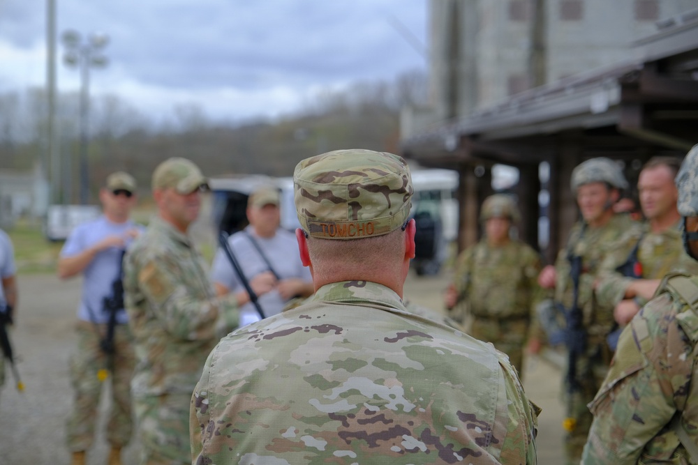 A group of military personnel in MultiCam/OCP camouflage uniforms are gathered outdoors at what appears to be a training facility or military installation. The central figure, whose name tape reads 'TOMCHO,' is facing away from the camera toward a group being addressed. The setting suggests a briefing or after-action review during a field training exercise.