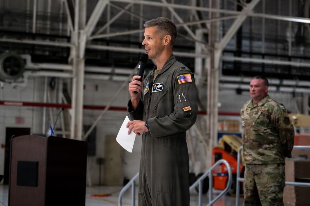 A military officer in an olive green flight suit is delivering a speech at a podium inside what appears to be an aircraft hangar. He is holding a microphone and papers while addressing an audience. A second military personnel member in camouflage uniform stands in the background.