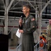 A military officer in an olive green flight suit is delivering a speech at a podium inside what appears to be an aircraft hangar. He is holding a microphone and papers while addressing an audience. A second military personnel member in camouflage uniform stands in the background.
