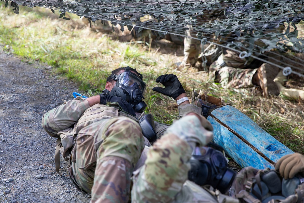 Two military personnel in camouflage uniforms are low-crawling under a camouflage net during what appears to be a training exercise. The lead soldier is wearing a full-face gas mask (CBRN protective mask) and tactical gloves while navigating under the obstacle. A blue object, likely a training prop or simulated IED/hazard marker, is visible alongside the soldiers.