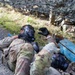 Two military personnel in camouflage uniforms are low-crawling under a camouflage net during what appears to be a training exercise. The lead soldier is wearing a full-face gas mask (CBRN protective mask) and tactical gloves while navigating under the obstacle. A blue object, likely a training prop or simulated IED/hazard marker, is visible alongside the soldiers.