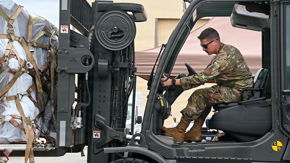 A military service member in MultiCam/OCP camouflage uniform is operating a military forklift, maneuvering what appears to be a palletized cargo load secured with straps and wrapped in white protective covering. The forklift bears a 'Cascade' brand attachment and displays capacity markings. The setting appears to be an outdoor military logistics or airfield operations area.