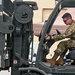 A military service member in MultiCam/OCP camouflage uniform is operating a military forklift, maneuvering what appears to be a palletized cargo load secured with straps and wrapped in white protective covering. The forklift bears a 'Cascade' brand attachment and displays capacity markings. The setting appears to be an outdoor military logistics or airfield operations area.
