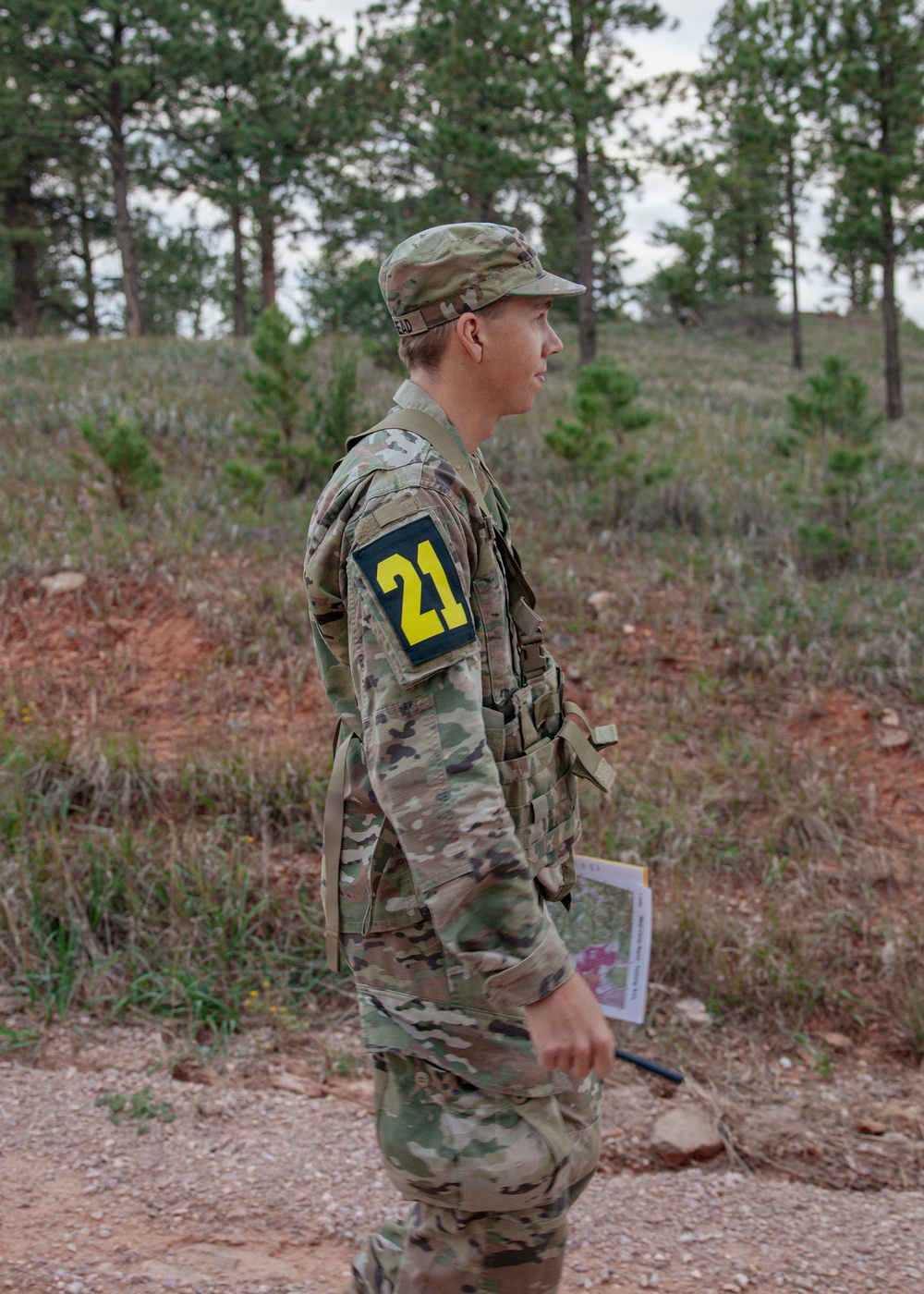 A soldier in US Army OCP (Operational Camouflage Pattern) uniform stands in a wooded, hilly terrain. The individual is wearing a numbered bib patch '21' on their left arm, suggesting participation in a military training exercise or competition. They are holding what appears to be a map or document, consistent with land navigation training.