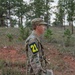 A soldier in US Army OCP (Operational Camouflage Pattern) uniform stands in a wooded, hilly terrain. The individual is wearing a numbered bib patch '21' on their left arm, suggesting participation in a military training exercise or competition. They are holding what appears to be a map or document, consistent with land navigation training.