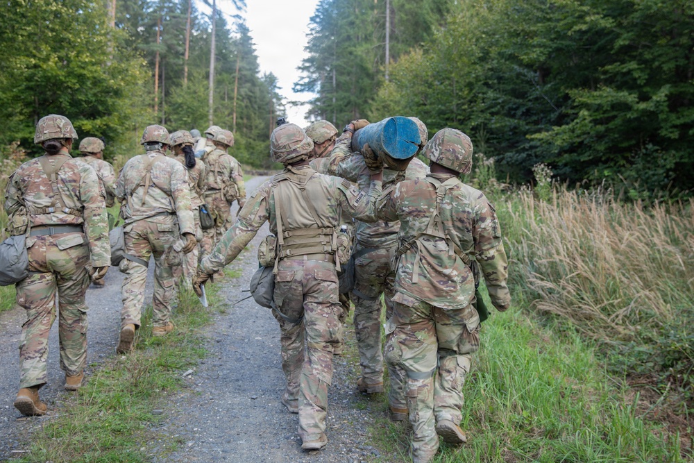 A group of military personnel in camouflage uniforms and helmets are marching along a forest trail. Two soldiers in the foreground are carrying a blue cylindrical object together, suggesting a team physical training or field exercise. The setting appears to be a wooded training area, likely in Central or Northern Europe based on the vegetation.