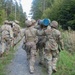 A group of military personnel in camouflage uniforms and helmets are marching along a forest trail. Two soldiers in the foreground are carrying a blue cylindrical object together, suggesting a team physical training or field exercise. The setting appears to be a wooded training area, likely in Central or Northern Europe based on the vegetation.