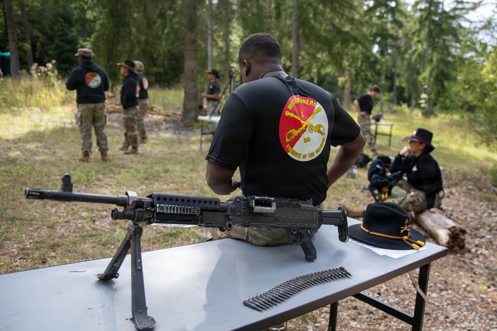 A group of individuals wearing matching black unit t-shirts are gathered at an outdoor range or training area surrounded by trees. In the foreground, a machine gun (appears to be an M240 or similar belt-fed weapon) is displayed on a folding table alongside a belt of ammunition and a cavalry hat with yellow cord. The setting appears to be a military unit event or demonstration, likely associated with a U.S. Army cavalry unit.