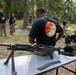 A group of individuals wearing matching black unit t-shirts are gathered at an outdoor range or training area surrounded by trees. In the foreground, a machine gun (appears to be an M240 or similar belt-fed weapon) is displayed on a folding table alongside a belt of ammunition and a cavalry hat with yellow cord. The setting appears to be a military unit event or demonstration, likely associated with a U.S. Army cavalry unit.