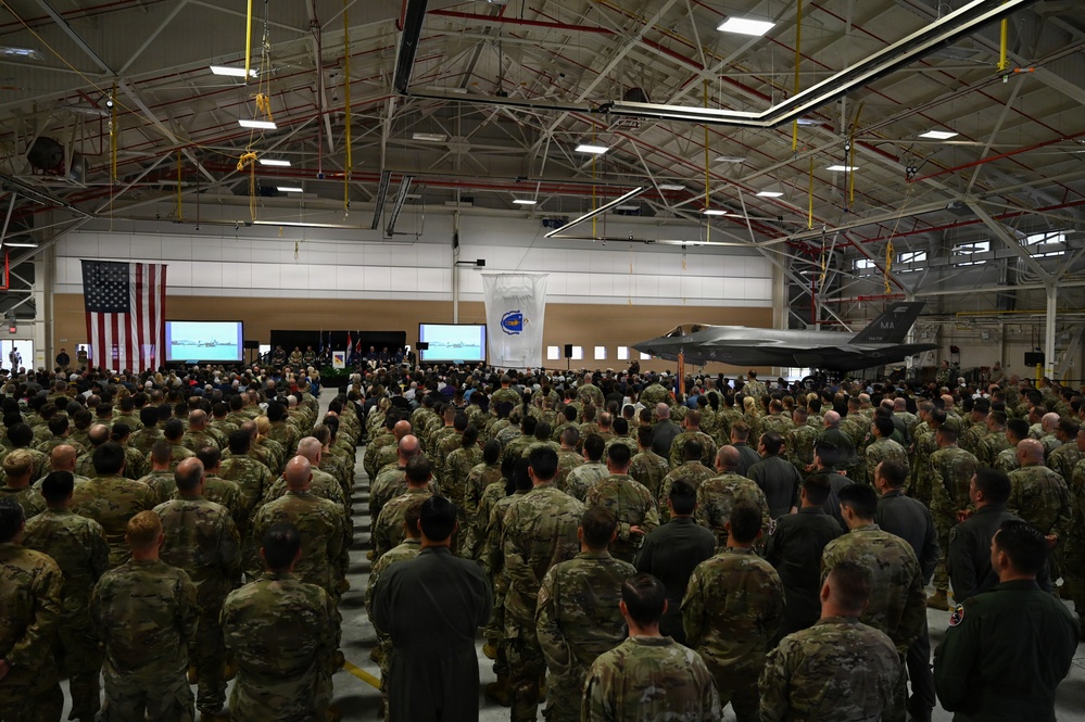 A large military ceremony is taking place inside an aircraft hangar, with hundreds of uniformed personnel standing in formation facing a stage or podium area. An F-35 fighter jet is visible on the right side of the hangar, and an American flag is prominently displayed on the left wall. Two large projection screens flank a central unit insignia banner, suggesting a formal change of command, activation, or award ceremony.