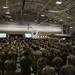 A large military ceremony is taking place inside an aircraft hangar, with hundreds of uniformed personnel standing in formation facing a stage or podium area. An F-35 fighter jet is visible on the right side of the hangar, and an American flag is prominently displayed on the left wall. Two large projection screens flank a central unit insignia banner, suggesting a formal change of command, activation, or award ceremony.