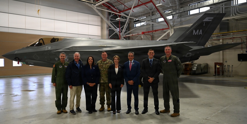 A group of eight individuals pose for a formal photograph in front of an F-35 fighter jet inside a military hangar. The aircraft displays 'MA' tail markings, indicating Massachusetts Air National Guard affiliation. The group includes military personnel in flight suits and camouflage uniforms alongside civilians in business attire, suggesting an official visit or ceremony.