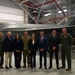 A group of eight individuals pose for a formal photograph in front of an F-35 fighter jet inside a military hangar. The aircraft displays 'MA' tail markings, indicating Massachusetts Air National Guard affiliation. The group includes military personnel in flight suits and camouflage uniforms alongside civilians in business attire, suggesting an official visit or ceremony.
