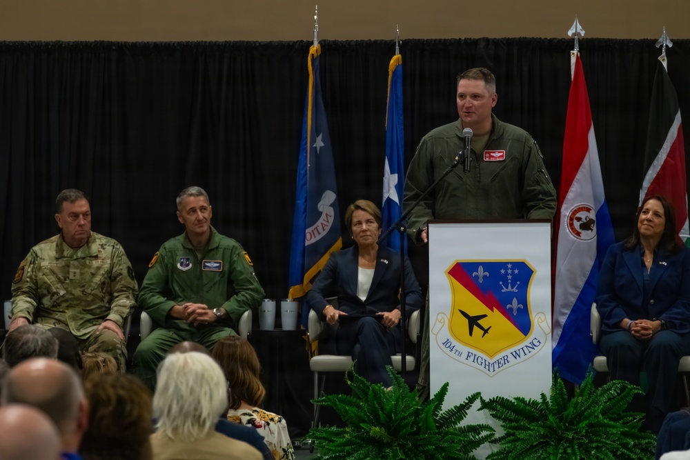 A formal military ceremony is taking place at the 104th Fighter Wing, with an officer in a green flight suit speaking at a podium bearing the unit's insignia. Several dignitaries and military personnel are seated on stage behind the speaker, with multiple flags displayed in the background. An audience is visible in the foreground attending the event.