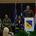 A formal military ceremony is taking place at the 104th Fighter Wing, with an officer in a green flight suit speaking at a podium bearing the unit's insignia. Several dignitaries and military personnel are seated on stage behind the speaker, with multiple flags displayed in the background. An audience is visible in the foreground attending the event.