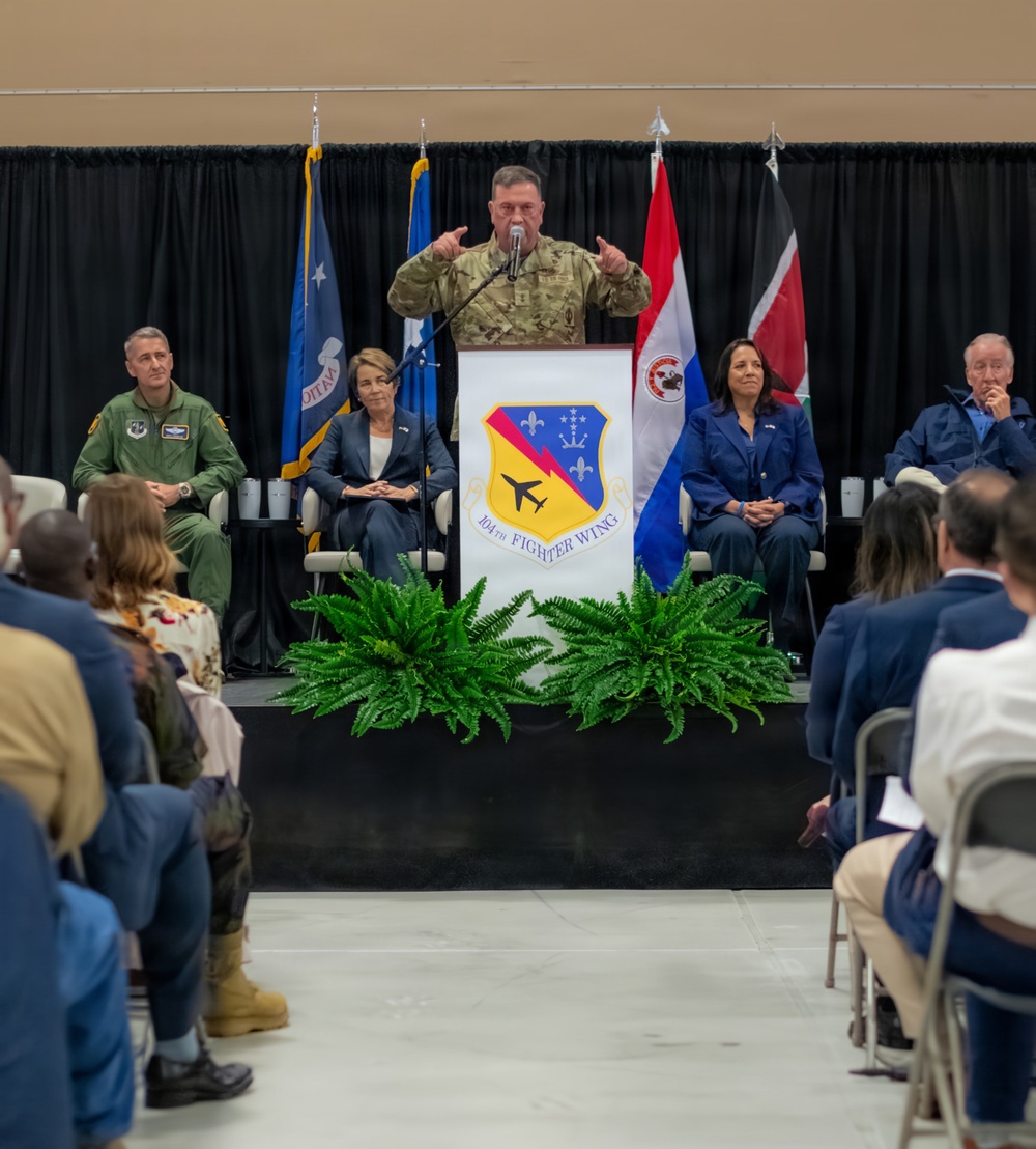 A formal military ceremony or press event is taking place at the 104th Fighter Wing, as indicated by the podium insignia. A military officer in camouflage uniform is speaking at the podium while several dignitaries, including civilians in business attire and a uniformed officer, are seated on stage. An audience is seated in folding chairs facing the stage.