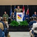 A formal military ceremony or press event is taking place at the 104th Fighter Wing, as indicated by the podium insignia. A military officer in camouflage uniform is speaking at the podium while several dignitaries, including civilians in business attire and a uniformed officer, are seated on stage. An audience is seated in folding chairs facing the stage.