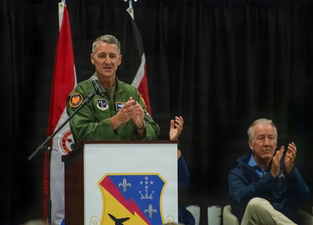 A U.S. Air Force officer in a green flight suit is delivering a speech at a podium during what appears to be an official military ceremony. The podium displays a unit shield/crest featuring fleur-de-lis, stars, and lightning bolt insignia. An older civilian man is seated in the background, applauding.