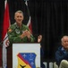 A U.S. Air Force officer in a green flight suit is delivering a speech at a podium during what appears to be an official military ceremony. The podium displays a unit shield/crest featuring fleur-de-lis, stars, and lightning bolt insignia. An older civilian man is seated in the background, applauding.