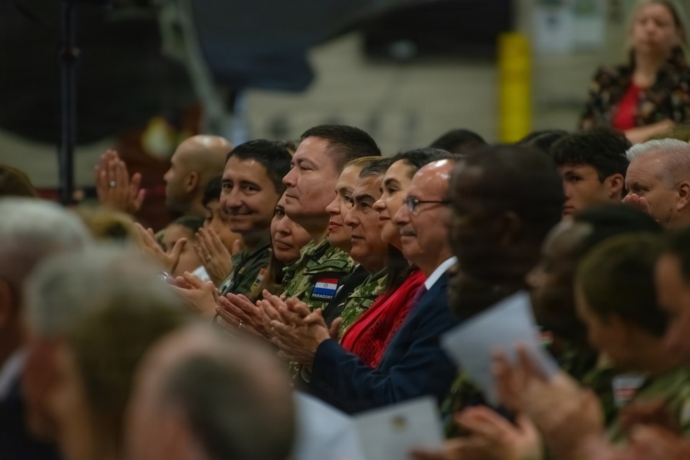 A diverse audience is seated and applauding at what appears to be a formal military ceremony or event. Several individuals are wearing military uniforms, including one with a visible 'PARAGUAY' name tape, alongside civilians in formal attire. The gathering appears to be multinational in nature, suggesting an international military or diplomatic event.