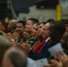 A diverse audience is seated and applauding at what appears to be a formal military ceremony or event. Several individuals are wearing military uniforms, including one with a visible 'PARAGUAY' name tape, alongside civilians in formal attire. The gathering appears to be multinational in nature, suggesting an international military or diplomatic event.