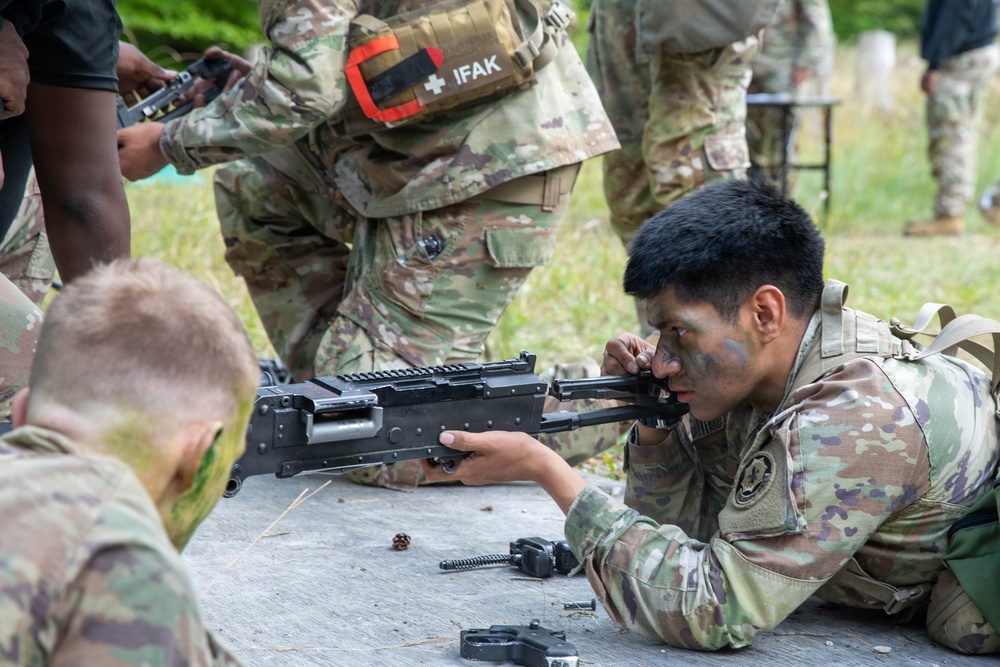Military personnel in Operational Camouflage Pattern (OCP) uniforms are engaged in a weapons maintenance or familiarization exercise outdoors. A soldier with camouflage face paint is closely inspecting or reassembling what appears to be an M249 Squad Automatic Weapon (SAW) on a flat surface. Disassembled weapon components are visible on the table, indicating a field-stripping or timed reassembly drill.