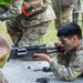 Military personnel in Operational Camouflage Pattern (OCP) uniforms are engaged in a weapons maintenance or familiarization exercise outdoors. A soldier with camouflage face paint is closely inspecting or reassembling what appears to be an M249 Squad Automatic Weapon (SAW) on a flat surface. Disassembled weapon components are visible on the table, indicating a field-stripping or timed reassembly drill.