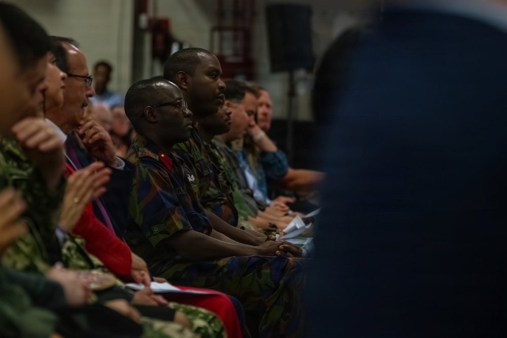 A group of military personnel and civilians are seated in rows, appearing to attend a formal briefing or ceremony in an indoor facility. At least two individuals in camouflage uniforms are prominently visible in the foreground, with one wearing what appears to be a red rank insignia. The audience appears attentive, suggesting an ongoing presentation or address.