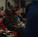 A group of military personnel and civilians are seated in rows, appearing to attend a formal briefing or ceremony in an indoor facility. At least two individuals in camouflage uniforms are prominently visible in the foreground, with one wearing what appears to be a red rank insignia. The audience appears attentive, suggesting an ongoing presentation or address.