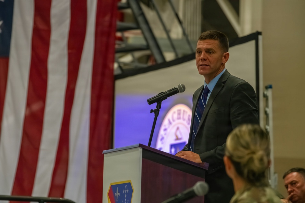 A man in a dark suit and blue shirt is speaking at a podium during what appears to be a military ceremony or official event. The podium displays a military unit insignia, and a large American flag is prominently displayed in the background. An audience including uniformed military personnel is visible in the foreground.