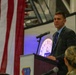 A man in a dark suit and blue shirt is speaking at a podium during what appears to be a military ceremony or official event. The podium displays a military unit insignia, and a large American flag is prominently displayed in the background. An audience including uniformed military personnel is visible in the foreground.