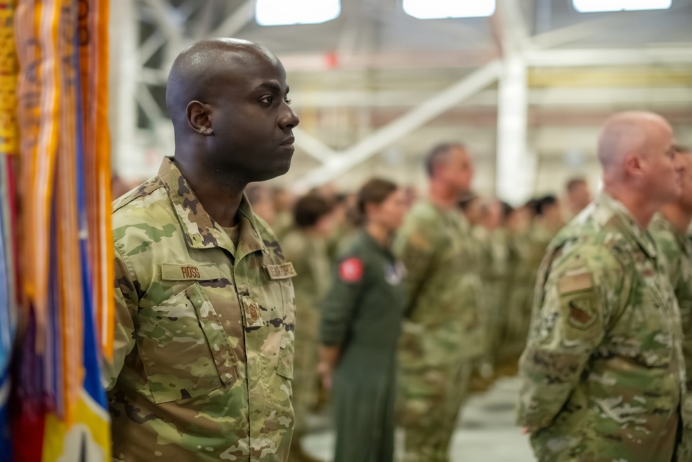 A military formation ceremony is taking place inside what appears to be an aircraft hangar. The foreground features a soldier with the name tape 'ROSS' wearing OCP (Operational Camouflage Pattern) uniform standing at attention. Multiple service members in various uniforms, including OCP and flight suits, are visible in formation in the background.