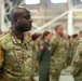 A military formation ceremony is taking place inside what appears to be an aircraft hangar. The foreground features a soldier with the name tape 'ROSS' wearing OCP (Operational Camouflage Pattern) uniform standing at attention. Multiple service members in various uniforms, including OCP and flight suits, are visible in formation in the background.