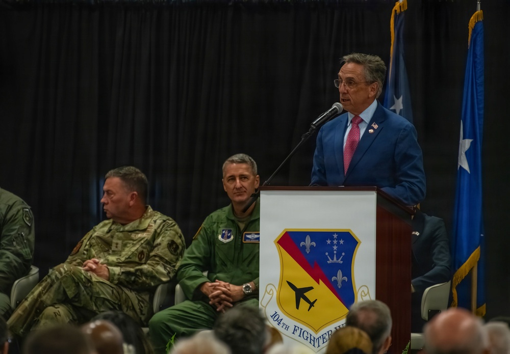 A formal military ceremony is taking place at the 104th Fighter Wing, as indicated by the unit insignia on the podium. A civilian man in a blue suit and pink tie is delivering a speech at the podium, while two military personnel — one in Army combat uniform and one in an Air Force flight suit — are seated behind him. Blue flags with stars are visible in the background.
