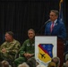 A formal military ceremony is taking place at the 104th Fighter Wing, as indicated by the unit insignia on the podium. A civilian man in a blue suit and pink tie is delivering a speech at the podium, while two military personnel — one in Army combat uniform and one in an Air Force flight suit — are seated behind him. Blue flags with stars are visible in the background.