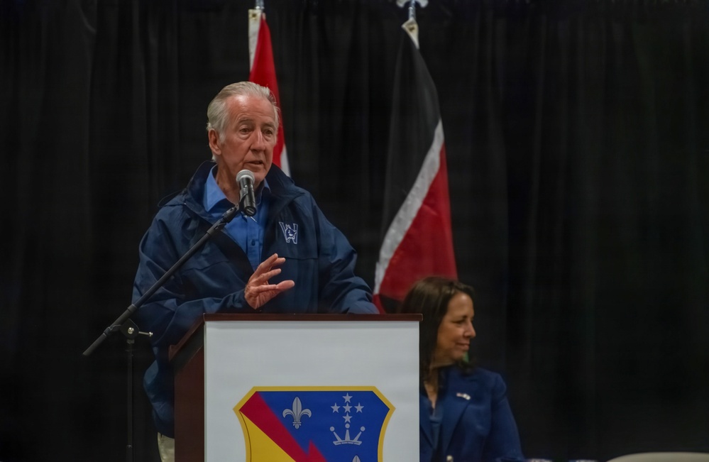 An elderly man in a blue jacket speaks at a podium equipped with a microphone at what appears to be an official or military-related event. A woman in a blue outfit is seated behind him to the right. The podium displays a military-style shield emblem, and flags are visible in the background.