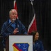An elderly man in a blue jacket speaks at a podium equipped with a microphone at what appears to be an official or military-related event. A woman in a blue outfit is seated behind him to the right. The podium displays a military-style shield emblem, and flags are visible in the background.