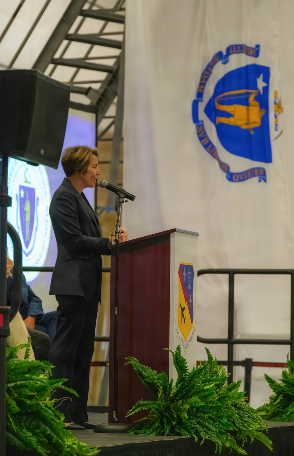 A person in a dark suit is speaking at a podium during what appears to be an official military or government ceremony. The setting includes a large banner displaying what appears to be a Massachusetts Air National Guard unit insignia, along with a projected Massachusetts state seal on a screen in the background. The event appears to be a formal ceremony held in a large hangar or similar facility.