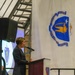 A person in a dark suit is speaking at a podium during what appears to be an official military or government ceremony. The setting includes a large banner displaying what appears to be a Massachusetts Air National Guard unit insignia, along with a projected Massachusetts state seal on a screen in the background. The event appears to be a formal ceremony held in a large hangar or similar facility.
