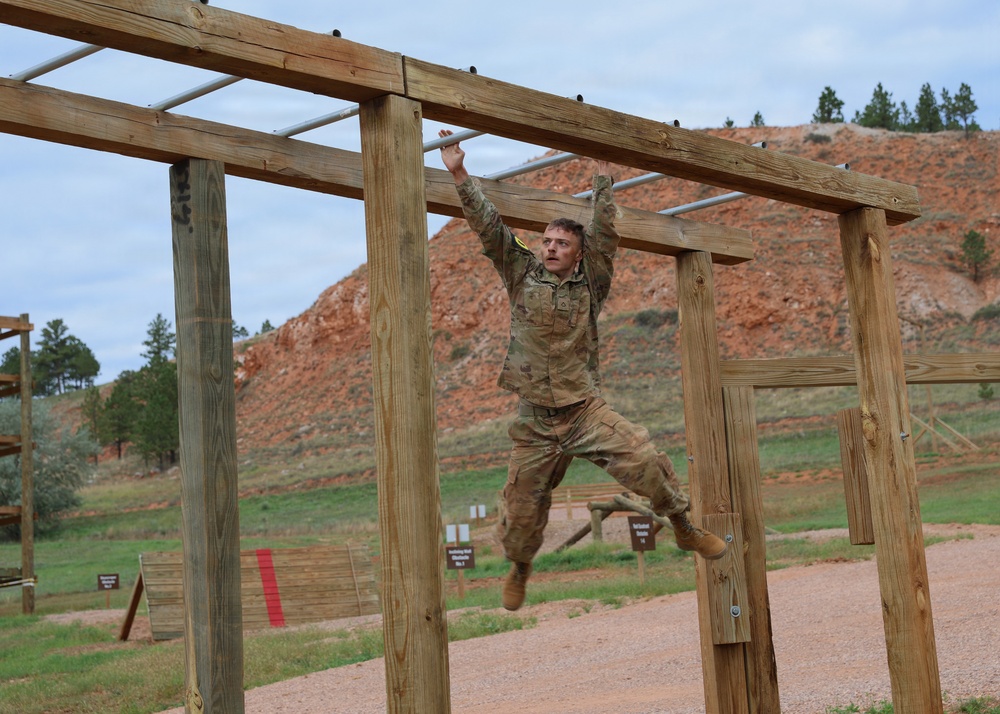A soldier in military camouflage (OCP/MultiCam) uniform is traversing monkey bars on an outdoor obstacle course. The setting appears to be a military training facility with red rock terrain and pine trees visible in the background. The soldier is mid-traverse, suspended by both hands from the horizontal bars.